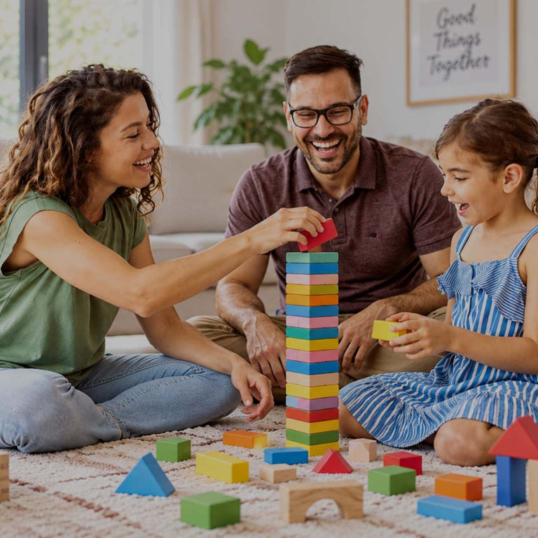A family of three plays with colorful wooden stacking blocks on a carpeted floor in a bright, modern living room. The mother, father, and daughter are smiling and engaged, with the daughter holding a yellow block and the tower being built high by the mother.