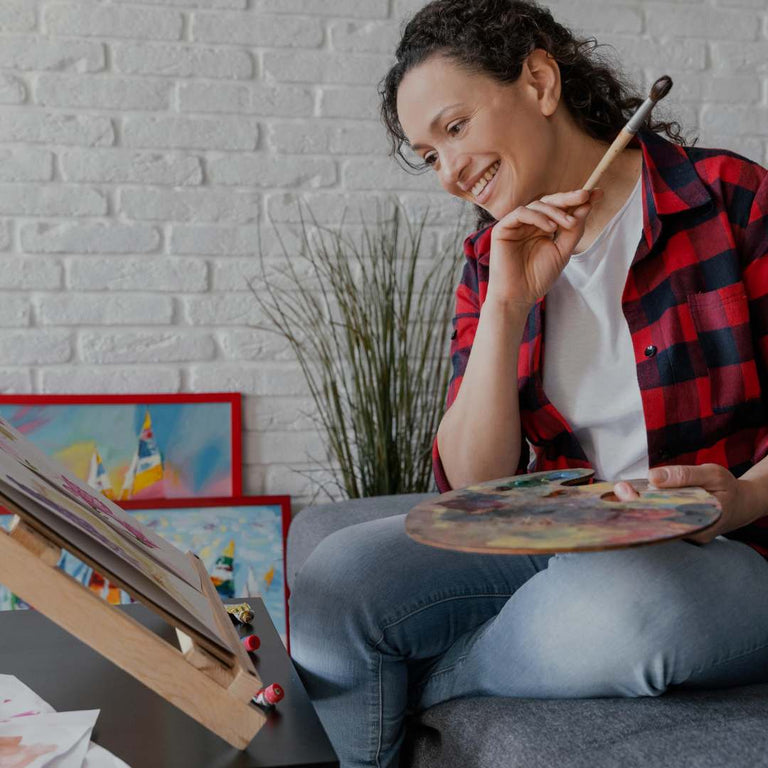 A woman with curly dark hair sits on a gray sofa in a living room setting, smiling and looking down at a paint palette she is holding. She is wearing a red and black plaid flannel shirt over a white t-shirt and blue jeans.