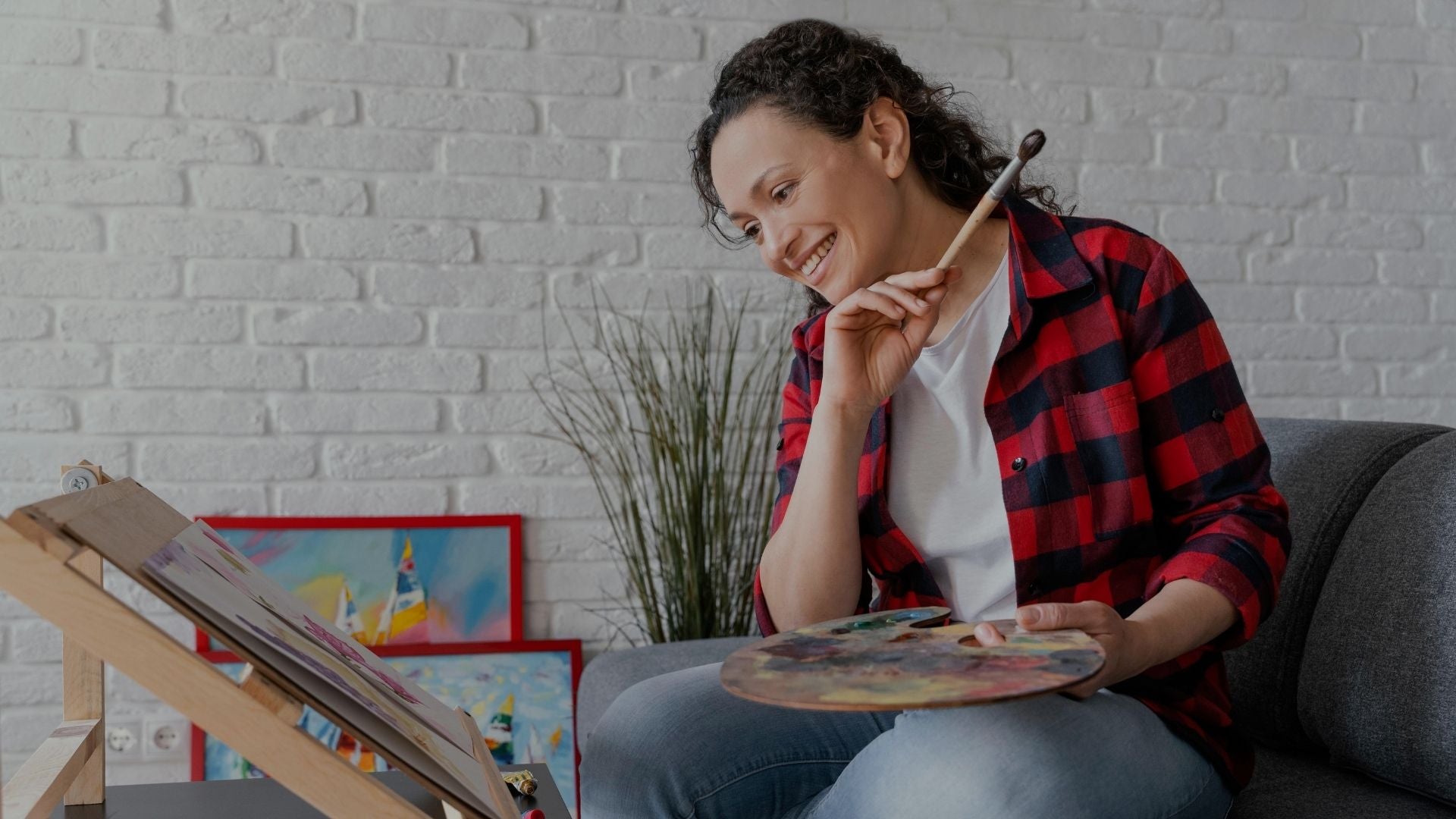 A woman with curly dark hair sits on a gray sofa in a living room setting, smiling and looking down at a paint palette she is holding. She is wearing a red and black plaid flannel shirt over a white t-shirt and blue jeans.